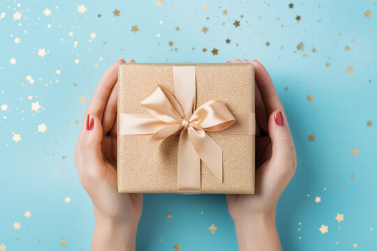 The photo shoot of a woman hands holding a wrapped gift box by paper and ribbon isolated on light blue background with metallic star glitter confetti or bokeh light. Generative AI.