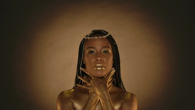Portrait of a seminude woman with golden skin and with golden metallic makeup on a brown background with circular light. The woman wraps her arms around her neck, touches the skin of her shoulders.