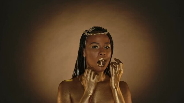 African American woman in Cleopatra style on a brown background with circular light. A woman with golden skin and jewelry on her head holds a golden chocolate candy with her lips, in her mouth.