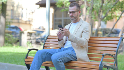 Middle Aged Man using Smartphone while Sitting Outdoor on a Bench