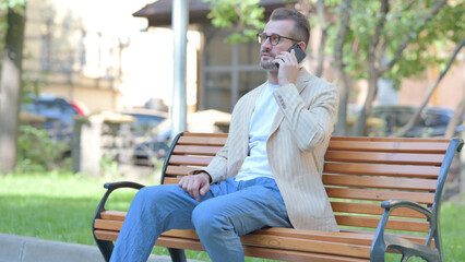 Fototapeta premium Middle Aged Man Talking on phone while Sitting Outdoor on a Bench