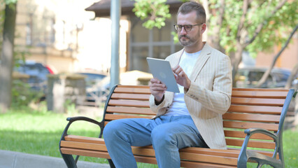 Middle Aged Man using Tablet while Sitting Outdoor on a Bench