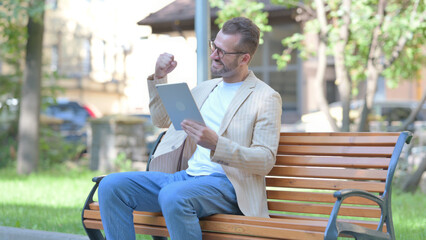 Fototapeta premium Middle Aged Man Celebrating Success on Tablet while Sitting Outdoor on a Bench