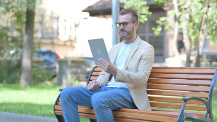 Middle Aged Man Doing Video Chat on Tablet while Sitting Outdoor on a Bench