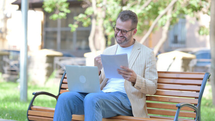 Middle Aged Man Celebrating while Working on Laptop and Documents Outdoor