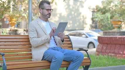 Fototapeta premium Modern Casual Man using Tablet while Sitting Outdoor on a Bench