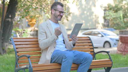 Modern Casual Man Celebrating Success on Tablet while Sitting Outdoor on a Bench