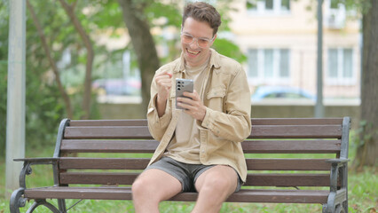 Casual Young Man Celebrating Success on Smartphone while Sitting Outdoor on a Bench