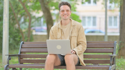 Fototapeta premium Casual Young Man Smiling at Camera while Working on Laptop Outdoor