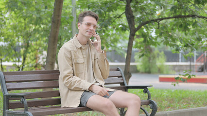 Fototapeta premium Modern Young Man Talking on phone while Sitting on Bench