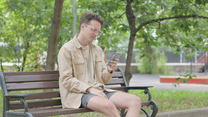 Fototapeta premium Modern Young Man Facing Loss on Smartphone while Sitting on Bench