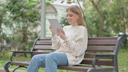 Obraz premium Casual Young Woman Celebrating on Tablet while Sitting Outdoor on a Bench