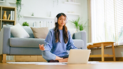 Happy young Asian woman practicing yoga and meditation at home sitting on floor in living room in lotus position and relaxing with closed eyes. Mindful meditation and wellbeing concept