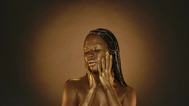 Portrait of a seminude African American woman with golden skin in the studio on a brown background with circular light. A woman rubs golden paint, liquid gold into the skin of her face.