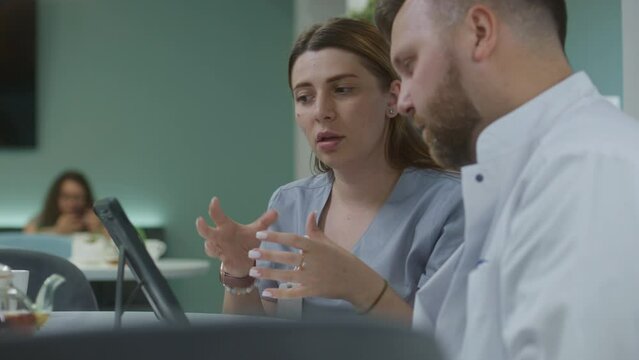 Female doctor sits in modern clinic cafe with male colleague, uses digital tablet. Professional medics talk, examine medical tests results, discuss diagnosis. Medical staff work in hospital canteen.