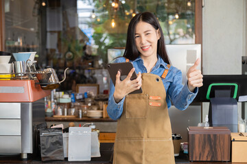 Happy Asian woman coffee business owner thumbs up hold tablet technology customer service order online delivery, young barista female standing in front of cafe counter and POS machine cash register