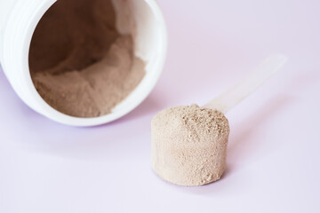 Brown protein powder in scoop and plastic jar on lilac background.