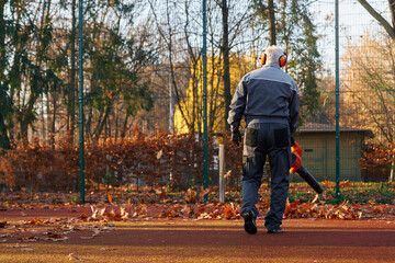 Gray haired man wearing earmuffs using portable leaf blower for cleaning up fenced park area. Back...
