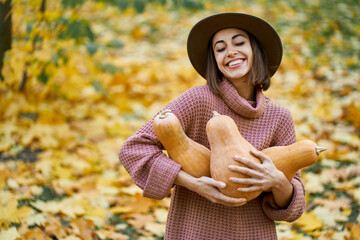 Pleased smiling lady in hat embracing big pumpkins surrounded orange leaves, enjoying autumnal time and seasonal harvest