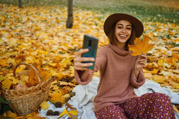 Autumn trendy style woman in hat and knit sweater captures the moment with selfie on smartphone at park with maple leafe in hand