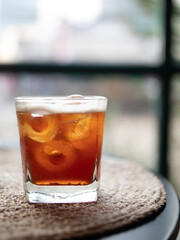 Iced tea is served in a small glass with a few leafy toppings placed on a placemat, isolated with a surrounding background.
