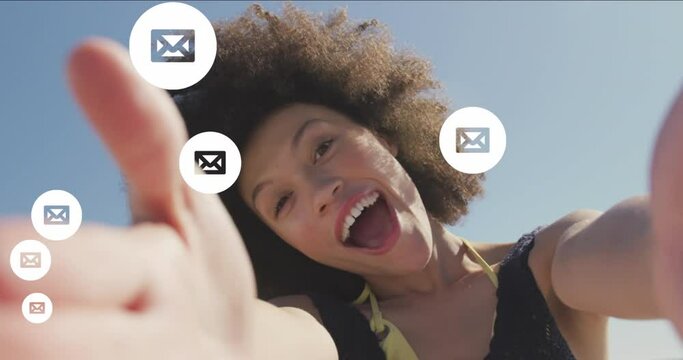 Animation of envelope icons over biracial woman holding camera and blowing kiss at beach