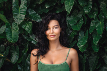 Natural beauty sport portrait woman with slim tanned body in stylish green sporty top posing near the wall of green exotic foliage and tropical leaves. High quality advertisement photo