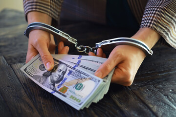 women's handcuffed hands hold dollars on a dark wooden background - obrazy, fototapety, plakaty