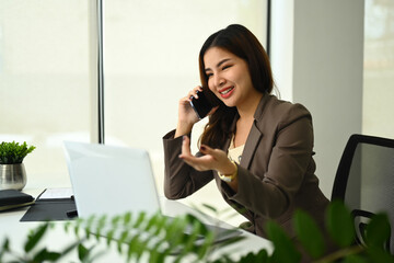 Successful businesswoman having pleasant phone conversation and using laptop in office