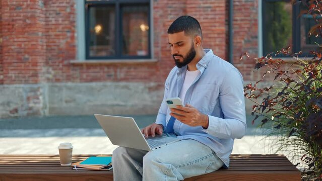 Serious indian man sitting on street bench outside with portable gadgets and revising information. Precise male in casual attire looking at smartphone and checking details on personal computer.