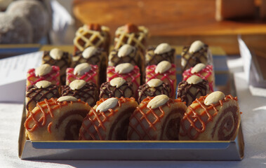 Pastry stand with variations of no-bake RAW cakes, roulade slices decorated with a lattice of chocolate or fruit cream and topped with blanched almonds, close-up without people at farmers market.
