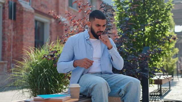 Indian man coughing and rubbing eyes because of feeling unwell while sitting on bench in downtown. Casually dressed guy feeling pain in breast, short of breath and nausea. Concept of health issues.