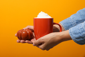 Red cup with coffee and decorative pumpkin in female hands on orange background