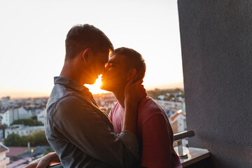 A young gay couple in love standing on a balcony overlooking the city at sunset and tenderly kissing