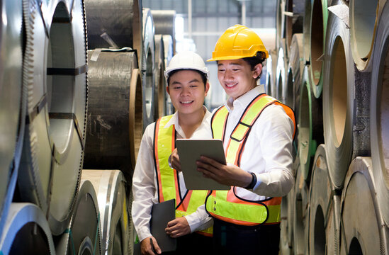 Two dedicated workers, adorned with protective hard hats, stand confidently in the industrial arena, surrounded by massive steel coils.