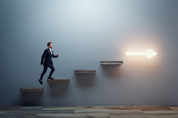 Side view of young man climbing illuminated arrow stairs to success on concrete wall background in...
