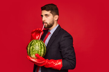 Stylish man in suit sitting at table in red rubber gloves, drinking watermelon, over red background. Concept of food, creativity, party. Pop art photography. Copy space for ad