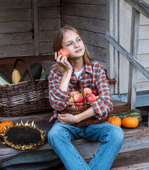 Girl is sitting on steps of an old village house with her face exposed to warm sunbeams, holding large basket with apples on her knees and eating apple. Ripe pumpkins and sunflower lie nearby. Harvest