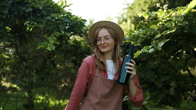 Happy young woman with glass bottle of red wine dancing and enjoying a day in vineyard winery countryside on sunny day. Toasting red wine glass at vineyard before sunset. High quality FullHD footage