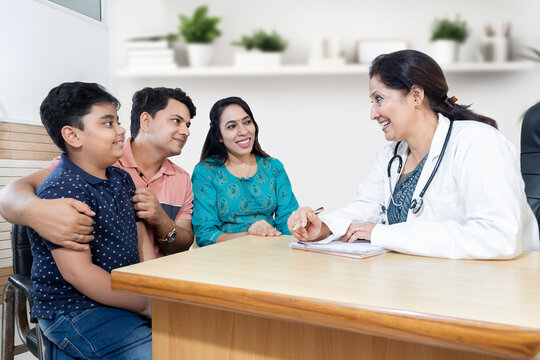 Young indian parents with their child boy patient consult female doctor in modern clinic. Regular pediatrician visit, Kid pediatric checkup in hospital, children healthcare and medical concept.