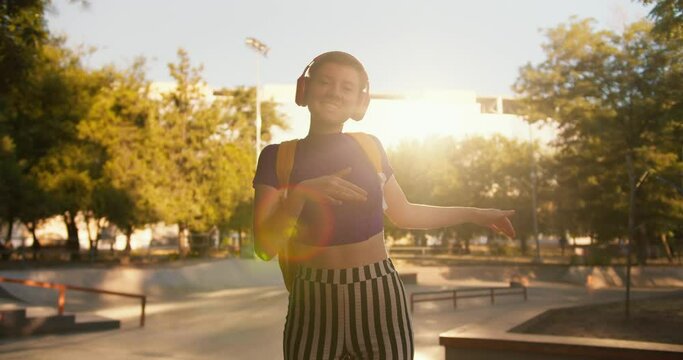 Portrait of a girl with a short haircut in a purple top, striped pants and red headphones is dancing on roller skates in a skatepark at sunset in summer