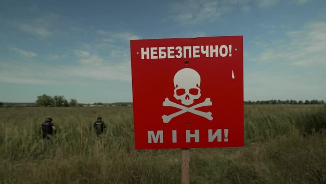 in the foreground a bright red sign with a skull and inscriptions about danger and mines, in the background a group of sappers working on demining the territory
mined field under a blue sky