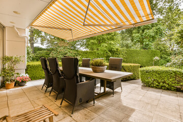 an outdoor dining area with table and chairs under a large yellow striped awning over the patio, surrounded by lush green trees - obrazy, fototapety, plakaty