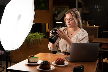 Smiling female food photographer with laptop computer and camera photographing cakes in cafe. Blogging and profession concept.