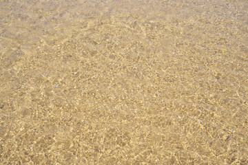 Top view of transparent clean water of sea ocean. Yellow sand seen through water. Sun rays reflecting on waves. Summer.