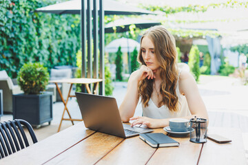 Young positive female freelancer sitting at a table with a laptop and working on a green terrace,...