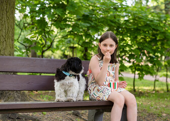 The junior girl is sitting on a bench in the park with her dog, enjoying ice cream from a cup.