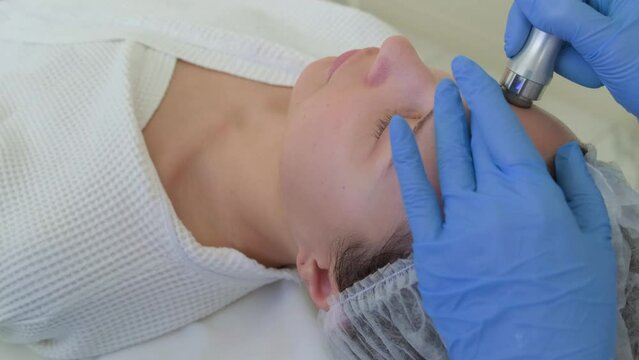 Professional dermatologist in medical gloves doing hydrafacial treatment on woman forehead. Closeup shot of vacuum peeling procedure at beauty spa.