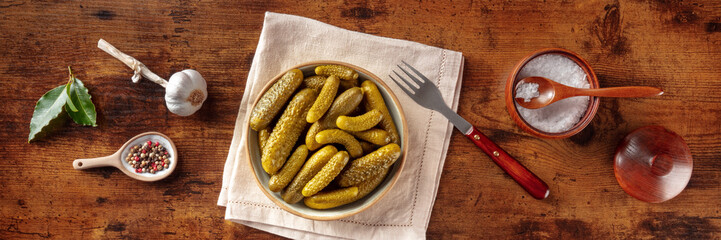 Pickled cucumbers with salt, pepper, garlic, and bay leaf panorama. Fermented food. Homemade canned gherkins. Overhead flat lay shot on a rustic wooden background, panoramic banner - obrazy, fototapety, plakaty