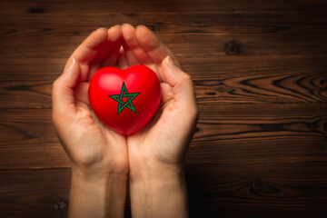 Women's hands hold and give a red heart in the shape of the Moroccan flag on a wooden background, a concept of support for earthquake victims